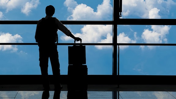 Mann mit Koffer sieht aus dem Fesnter eines Flughafens