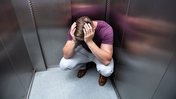 Ein Mann kauert in einem Aufzug mit Händen am Kopf