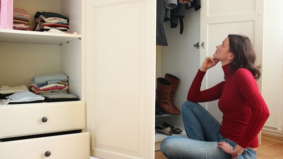 Eine junge Frau sitzt vor ihrem übervollen Kleiderschrank.