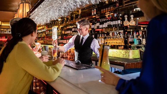 Frauen sitzen an einer Bar, ein Barkeeper mixt ein Getränk. 