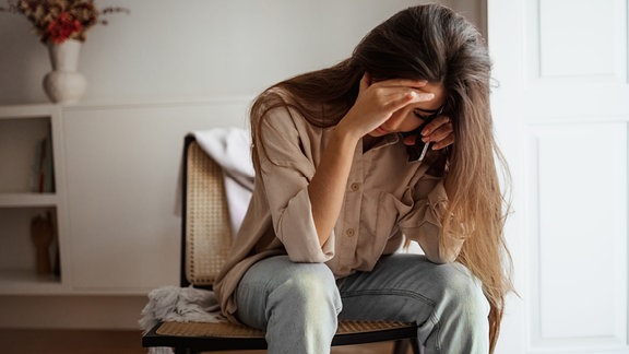 Symbolbild: eine junge Frau auf einem Stuhl hält sich den Kopf und ein Smartphone ans Ohr.