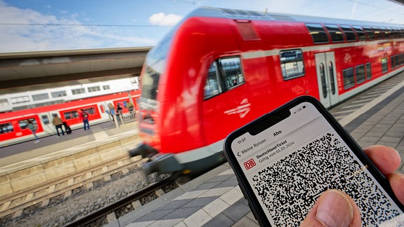 Jemand hält ein Smartphone in die Kamera, auf dem ein Deutschlandticket angezeigt wird. Im Hintergrund fährt ein Zug in den Bahnhof ein.