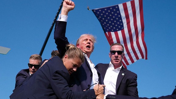 Donald Trump reckt blutverschmiert und abgedeckt von seinen Personenschützern die Faust in die Luft