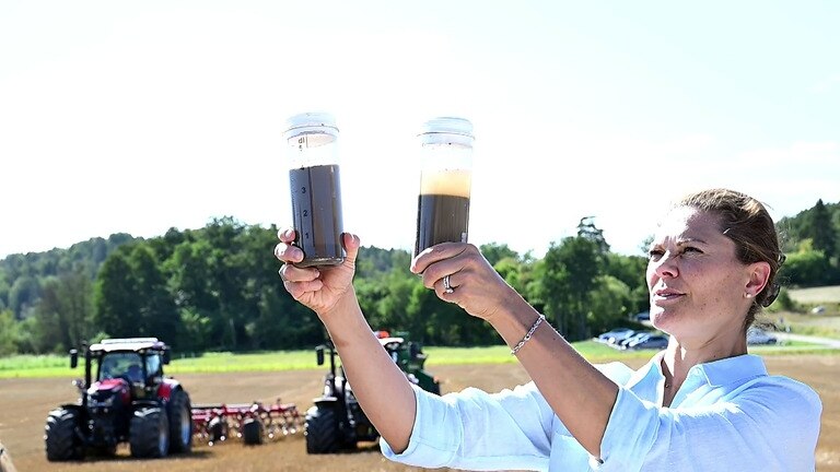 Victoria von Schweden hält, auf einem Acker stehend, zwei Bodenproben in den Händen