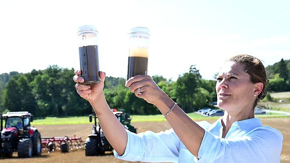 Victoria von Schweden hält, auf einem Acker stehend, zwei Bodenproben in den Händen