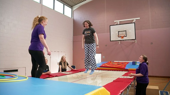 Eine junge Frau auf einem Trampolin, um sie herum stehen andere Frauen