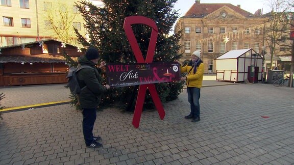 Zwei Personen stehen vor einer mannshohen Aidsschleife vor einem Weihnachtsbaum.