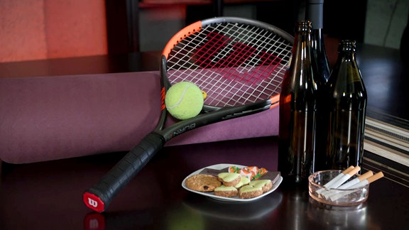 Auf einem Tisch liegen süßigkeiten, Zigaretten, Bierflaschen und Tennisschläger.