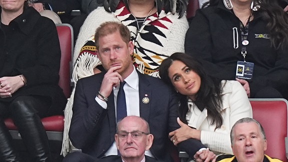 Harry und Meghan sitzen mit anderen Menschen auf einer Tribüne.