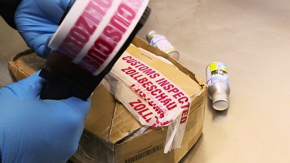 Eine Person verschließt ein Paket mit einem Klebeband auf dem "Zollbeschau" zu lesen ist.