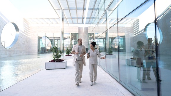Zwei Personen in weißer Medizin-Kleidung laufen durch ein verglastes Gebäude.