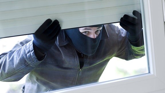 Maskierter Mann hebt Rollladen an und sieht durch ein Fenster