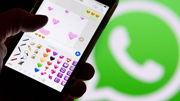 Ein Smartphone mit Emojis aus WhatsApp vor dem WhatsApp-Logo. 