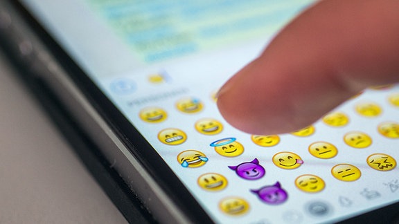 Eine Frau tippt auf das Display eines Smartphones, auf dem zahlreiche Emojis in der Nachrichten-App "Whatsapp" zu sehen sind.