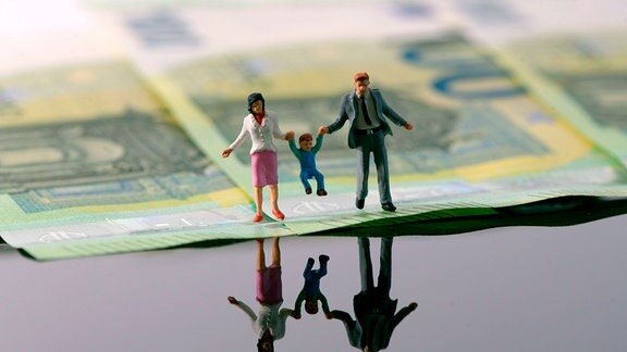 Symbolbild Finanzen: Kleine Puppen stehen auf Geldscheinen und schauen auf eine spiegelnde Fläche. Sie stellen eine Familie mit Kleinkind am See dar.