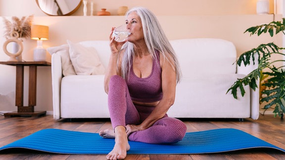 Frau sitzt auf einer Yogamatte und trinkt ein Glas Wasser.