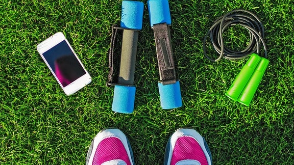 Weibliche Beine mit Turnschuhen auf grünem Gras mit Hanteln, Telefon und Springseil. Aktiver Lebensstil 