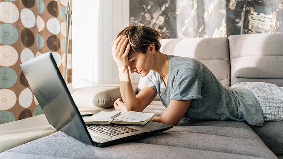 Eine junge Frau auf einem Sofa vor einem Laptop hält sich den Kopf
