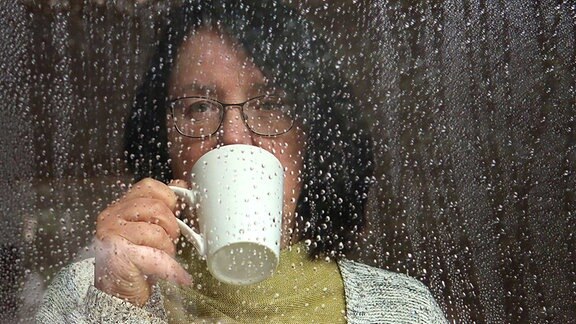 Eine Frau mit einer Tasse vor dem Mund schaut durch ein regennasses Fenster