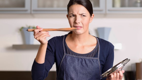 Frau mit Kochlöffel probiert ungenießbares Essen.