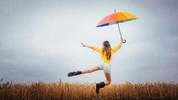 Eine Frau mit Regenschirm springt in die Luft. Dahinter ein Kornfeld.