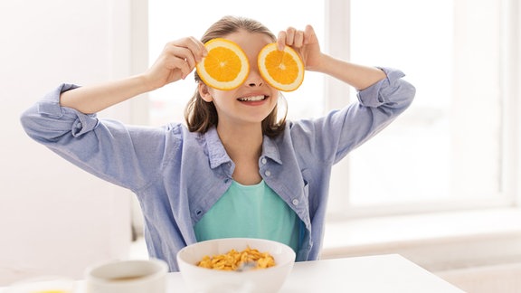 Ein Mädchen hält sich Orangenscheiben vor die Augen beim Frühstück zu Hause.