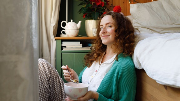 Frau sitzt mit einer Schüssel Essen zu Hause am Bett.