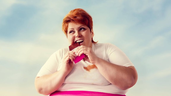 Übergewichtige Frau sitzt auf einem Dach und beißt in eine Schokoladentafel.