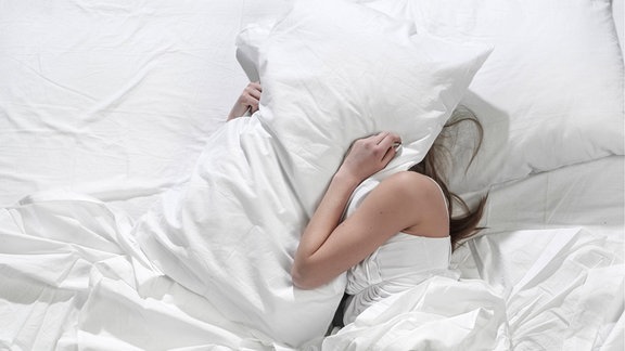 Eine Person liegt im Bett und hält sich ein Kissen über den Kopf.