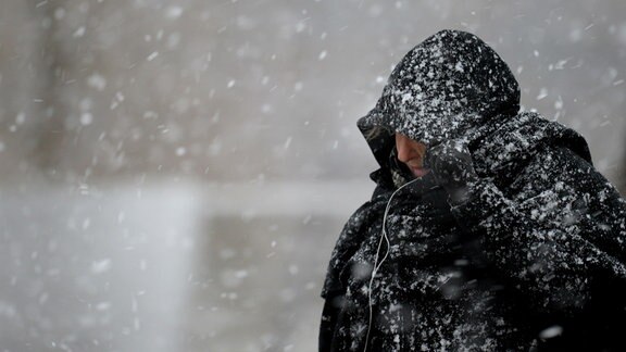Eine Passant*in zieht sich in einem Schneesturm die Kapuze ihrer Jacke ins Gesicht.