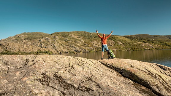 Ein Mann steht mit nach oben gestreckten Armen auf einem Felsen neben einem See.