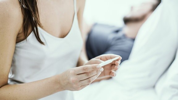 Junge Frau sitzt auf Bett und hält Schwangerschaftstest