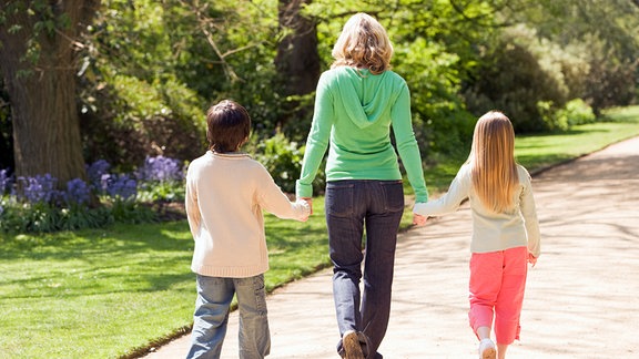 EIne Mutter spaziert mit ihren beiden Kindern durch einen Park.