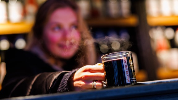 Eine Mitarbeiterin eines Glühweinstandes auf dem Weihnachtsmarkt hält eine dampfende Tasse Glühwein in der Hand. 