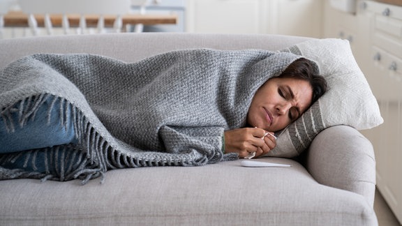 Eine Frau liegt, in eine Decke eingewickelt, auf dem Sofa.