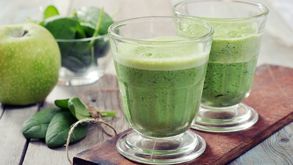 Ein Smoothie aus Apfel und Spinat steht auf einem Tisch. Die frischen Zutaten liegen daneben.