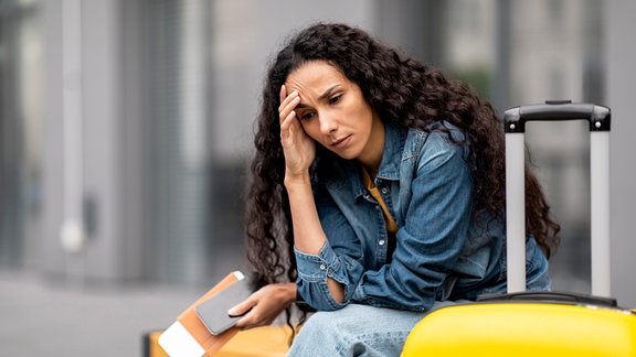Eine Frau sitzt neben ihrem Koffer und fasst sich an den Kopf.