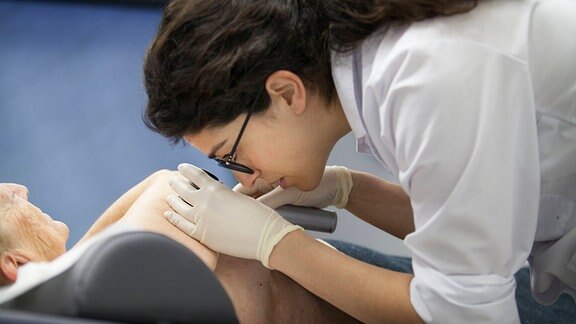Eine Dermatologin untersucht die Haut eines Patienten mit einer Lupe.