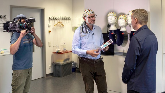 Vor der MDR-Kamera im Gespräch: Eckart von Hirschhausen (Mitte) mit einer EEG-Haube und Informatiker Dr. Christoph Reichert. Er hat eine Methode entwickelt, die bald Locked-in-Patienten zugutekommen soll.