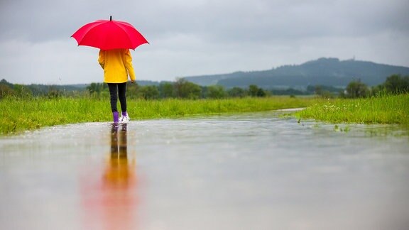 Eine Spaziergängerin in Gummistiefeln läuft im Regen auf einem überfluteten Weg entlang.