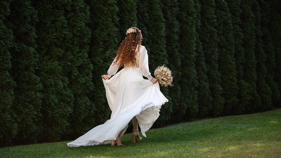 Eine Frau in einem Hochzeitskleid läuft an einer Hecke entlang