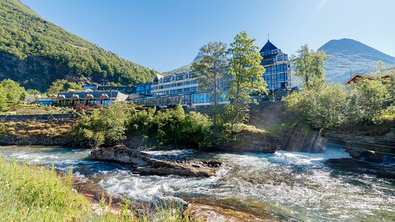 Ein Gebäude mit Glasfassade in Berglandschaft, im Vordergrund ein Fluss.