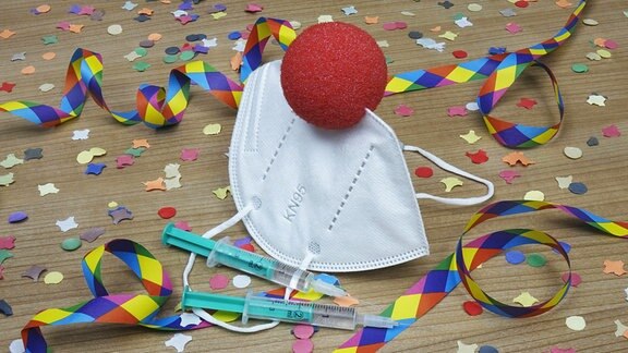 .Zwischen Papierschlangen und Konfetti liegt eine Mundschutzmaske mit einer roten Clownnase.