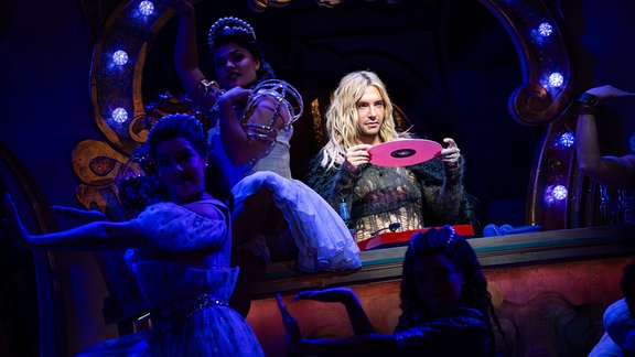 Bill Kaulitz steht neben Tänzerinnen auf einer Bühne und hält eine pinkfarbene Schallplatte hoch.