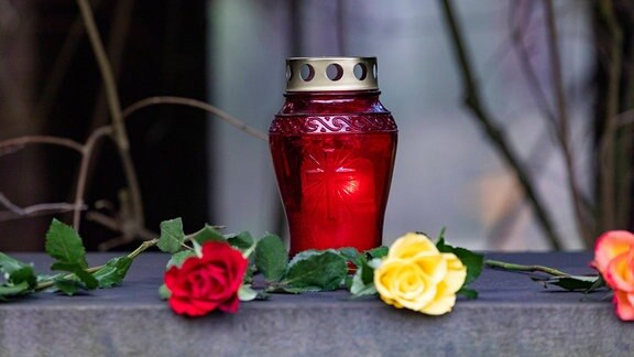 Eine rote Grabkerze auf einem Grab mit einer roten und gelben Rose im Vordergrund