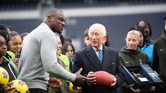 König Charles III. erhält einen NFL-Football, während Spurs-Frauenkapitänin Beth England bei einem Besuch im Tottenham Hotspur Stadium in London zuschaut.