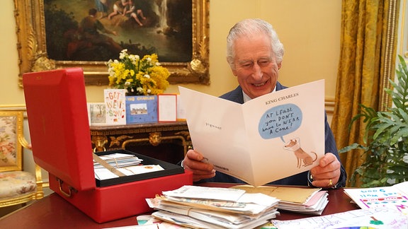 Der britische König Charles III. sitzt an einem Schreibtisch mit einer roten Box. Er liest eine Karte mit Genesungswünschen.