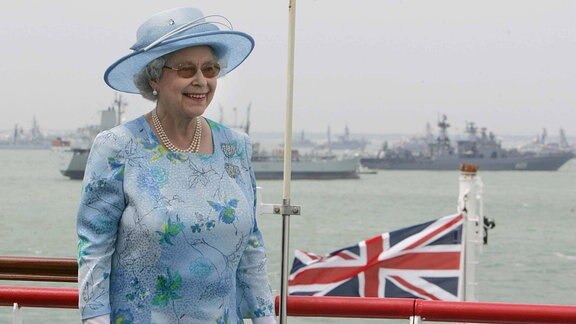 Die Queen vor Schiffen am Meer. Im Hintergrund der Union Jack.
