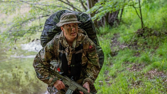 Eine Person in Uniform mit Tarnfarben und einem großen Rucksack läuft mit einer Waffe durch einen Wald.