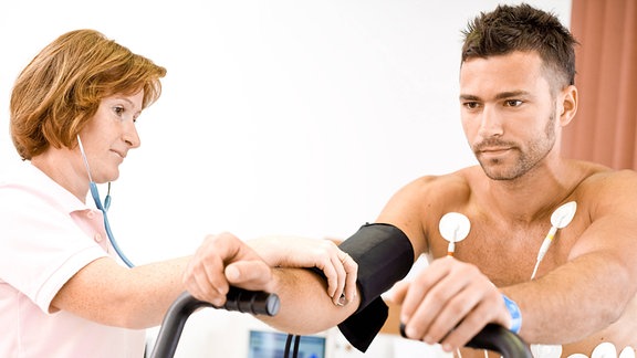 Ein Mann auf einem Fitnessgerät wird von einer Ärztin untersucht